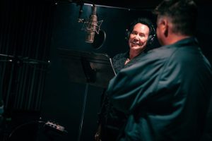 Wayne Newton in black jacket and headphones recording vocals for the song 'This Is America' next to a microphone